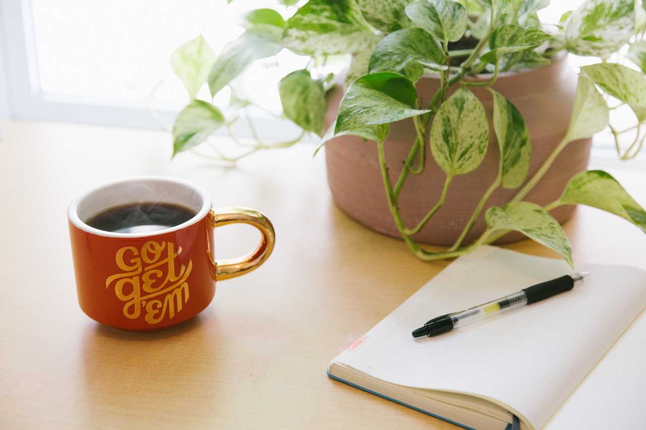 cup of coffee with a good day wish next to a plant and a notebook over a wooden table