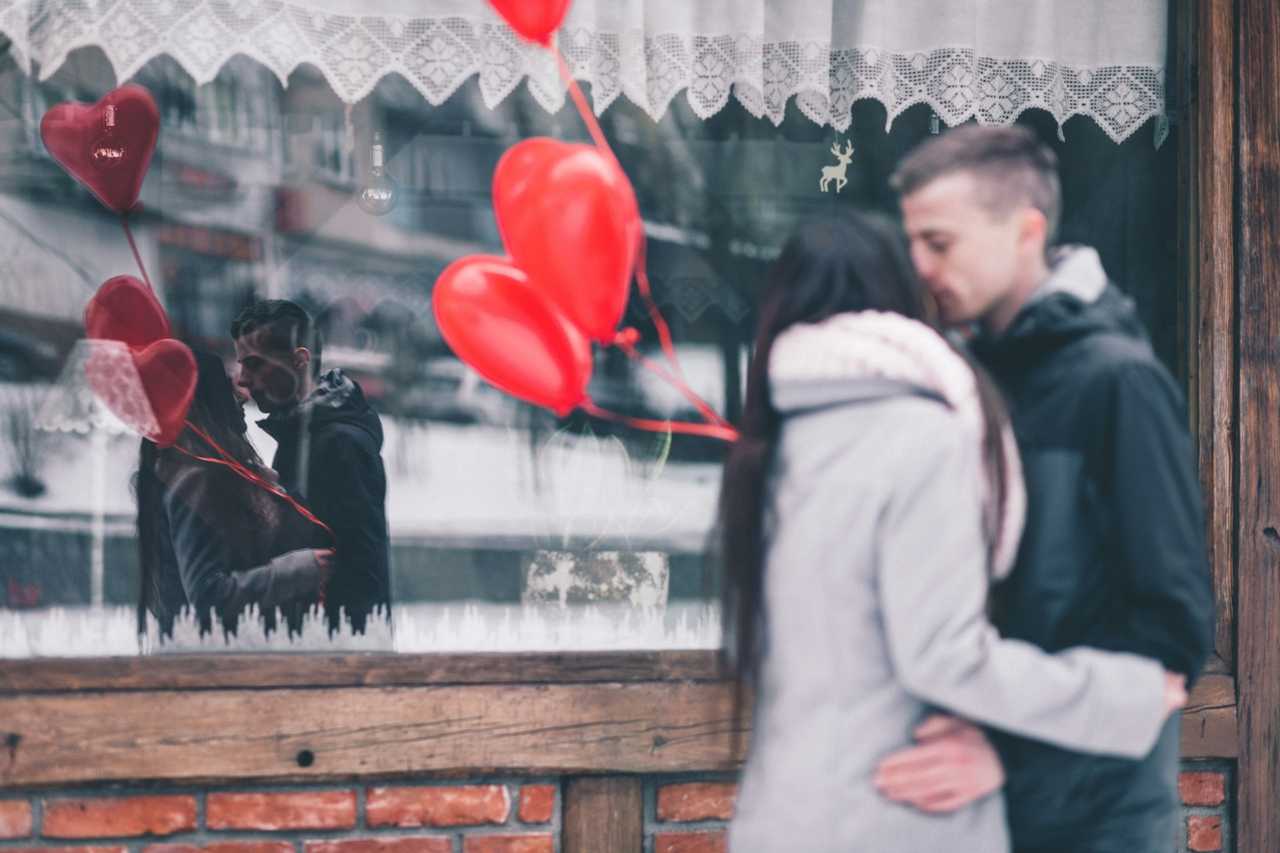 man and woman kissing on first date wearing coats in front of a store and holding red air balloons